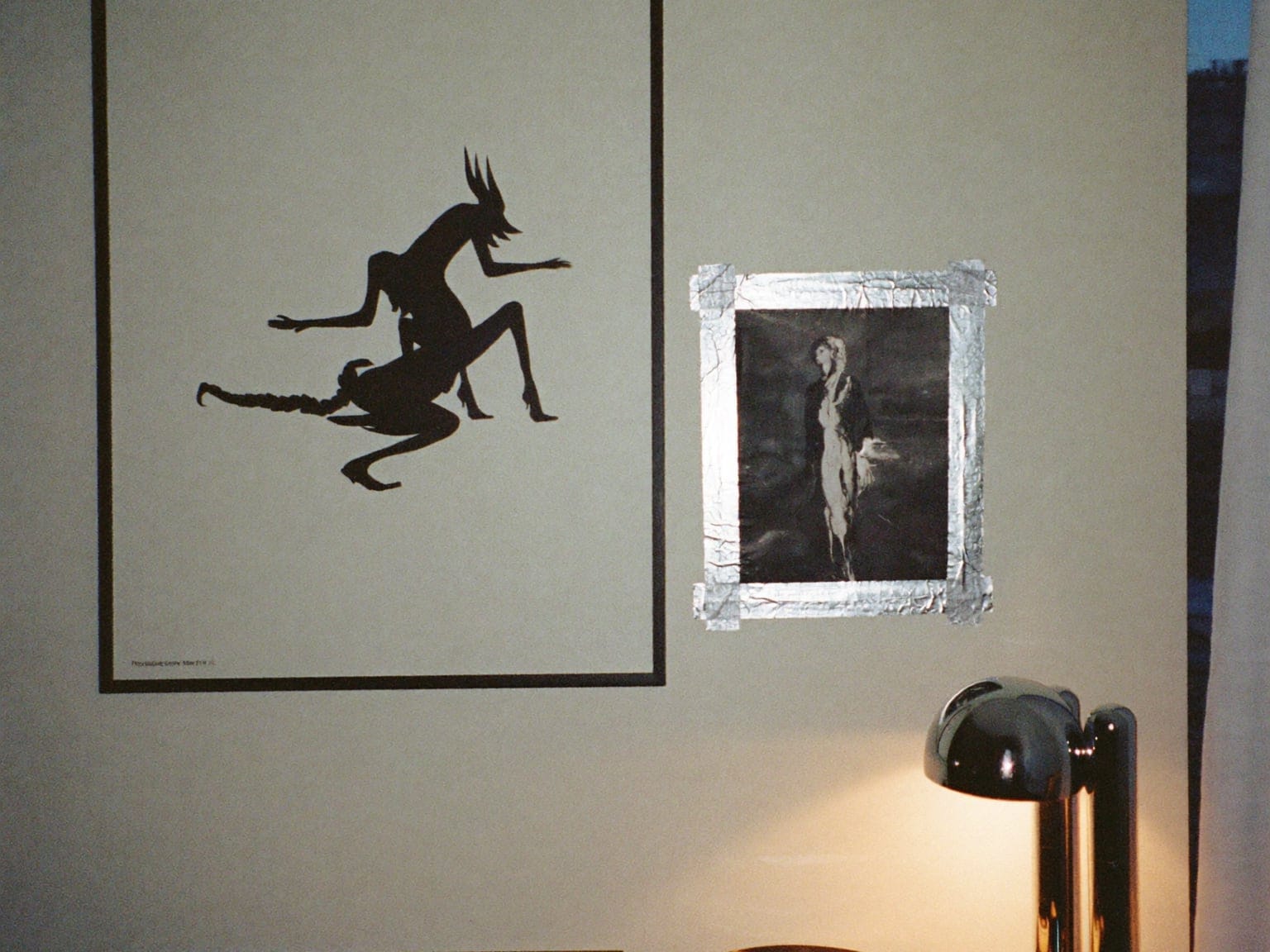 A dim room: a framed black-and-white poster, a smaller print taped up with silver tape, a chrome lamp