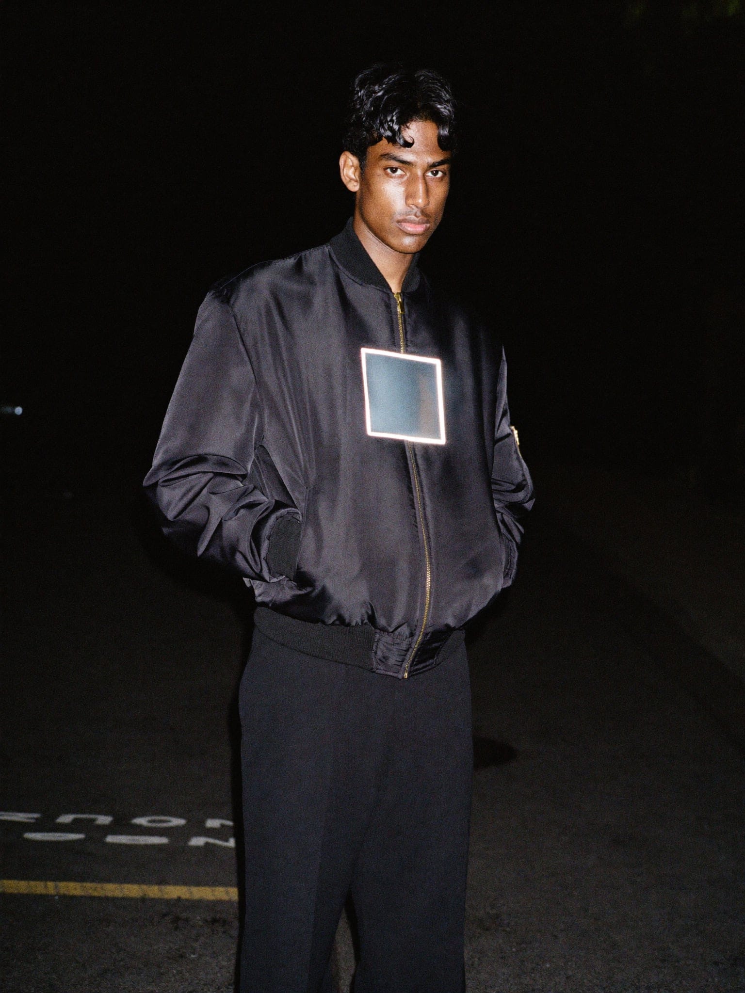 Model at night in a black satin bomber, the chest panel glowing white under flash