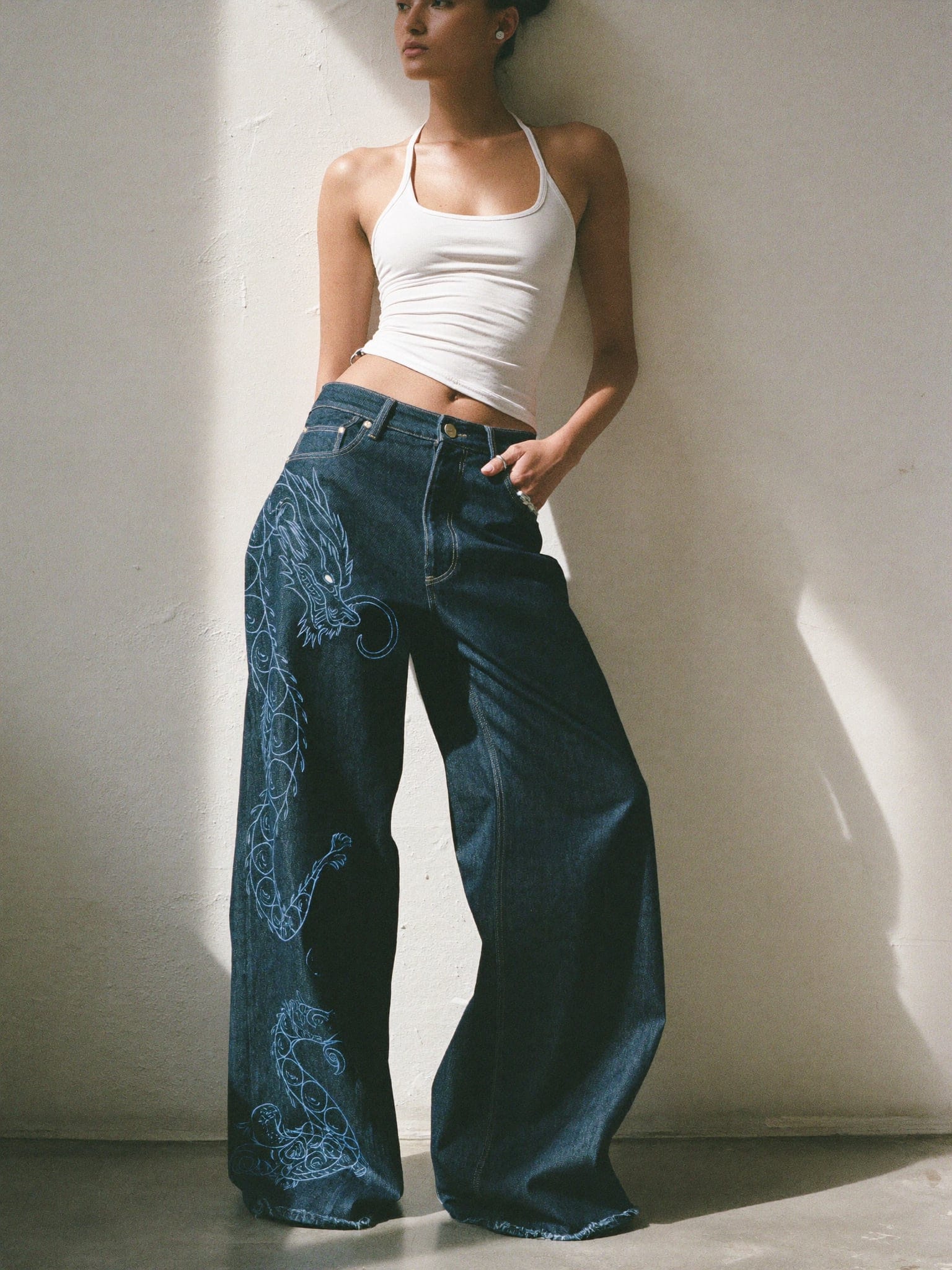 Model leaning on a sunlit wall in extra-wide indigo jeans with a pale embroidered dragon down the left leg