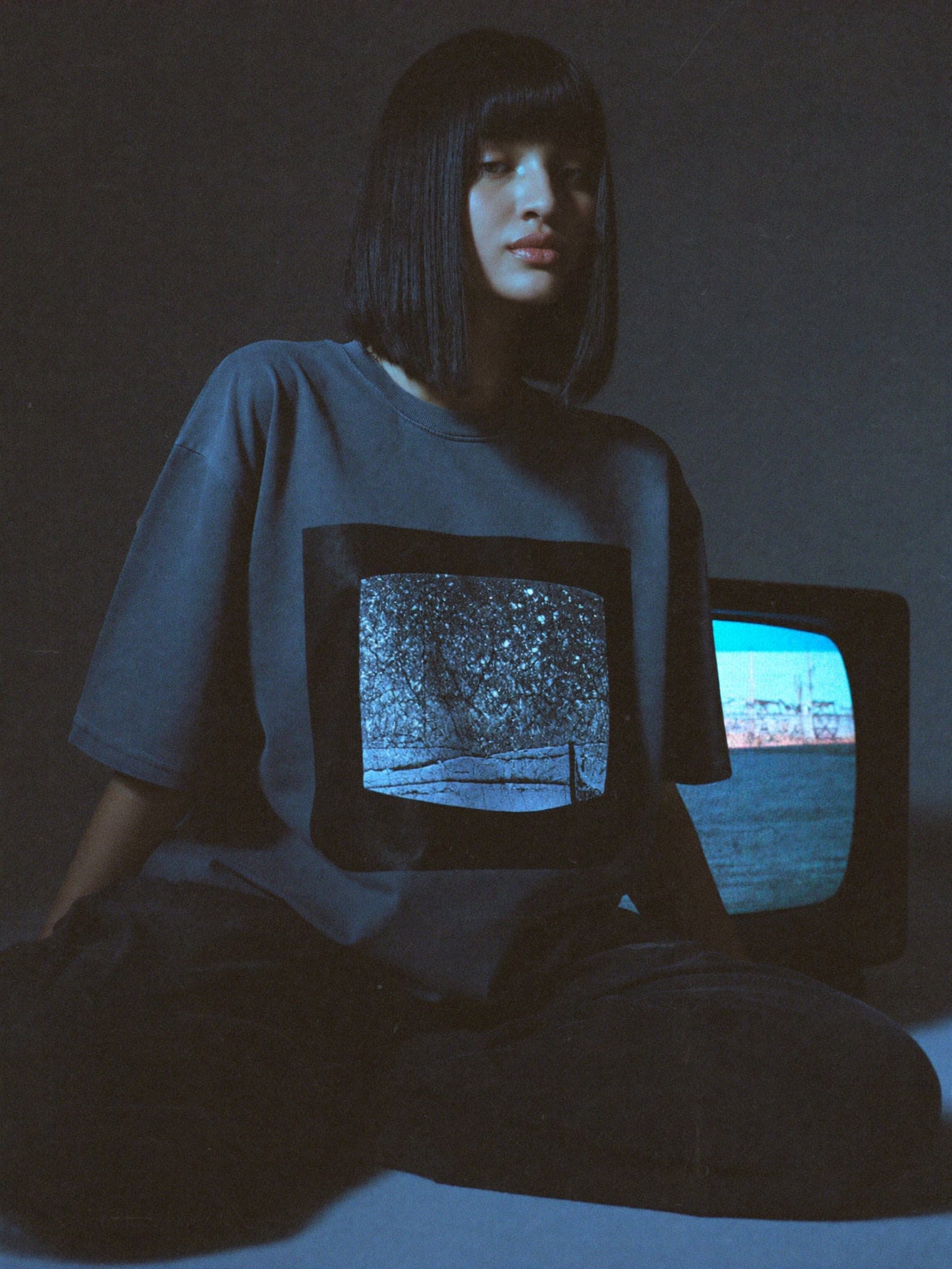 Model sitting in a dark room beside an old television, wearing a charcoal tee with a framed static print